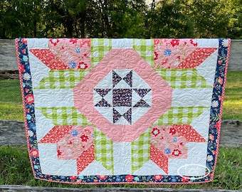 Op de afbeelding: Een handgemaakte quilt met een centraal sterrenpatroon in marineblauw en wit, omgeven door een roze ruit. De quilt heeft bloemen- en geometrische patronen in roze, rood, groen en wit, met een marineblauwe bloemenrand.