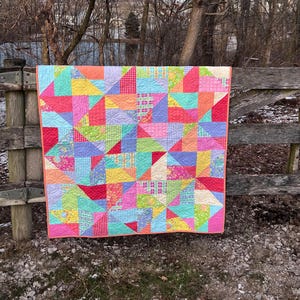 Puede incluir: Un edredón de patchwork colorido con formas geométricas en tonos de rosa, rojo, azul, verde y amarillo. El edredón tiene un borde rosa y se exhibe al aire libre.