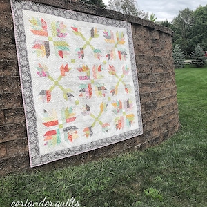 May include: A colorful quilt with a white background and a floral pattern in shades of red, orange, green, and pink. The quilt is bordered with a gray floral pattern and is displayed outdoors. The text "coriander quilts" is in the bottom left corner.