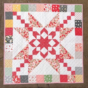 May include: A square patchwork quilt featuring a central star pattern in red and white, surrounded by a border of multi-colored floral and geometric fabric squares. The entire quilt is finished with a thin red and white checkered fabric binding.