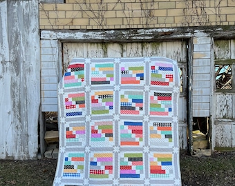 May include: A colorful patchwork quilt with a gray border, featuring rectangular blocks in various colors and patterns. The quilt is displayed outdoors against a weathered building. The quilt has a star pattern in the corners of each block.