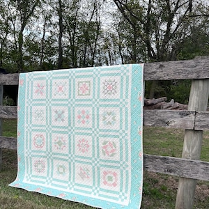 May include: A handmade quilt with a light blue border and a white center featuring a grid pattern with floral and geometric designs in pink and green. The quilt is draped over a wooden fence in an outdoor setting.