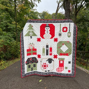 Op de afbeelding: Een kerstquilt met een witte achtergrond en diverse applicatieontwerpen. De quilt toont een kerstboom, sneeuwpop, cadeaus, kaarsen, krans, schaap en kous. De quilt heeft een zwart-grijze rand en een rode rand.