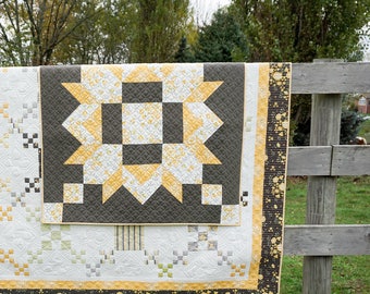 May include: A handmade quilt with a starburst design in shades of gray, yellow, and white. The quilt is draped over a wooden fence, showcasing its intricate patterns and textures. The quilt's border features a black and yellow speckled design.