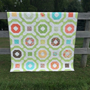 May include: A handmade quilt with a geometric pattern in shades of green, white, orange, and teal. The quilt is draped over a wooden fence, showcasing its intricate design and craftsmanship. The quilt features a repeating pattern of octagons and squares.