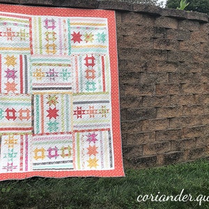 Op de afbeelding: Een kleurrijke patchworkquilt met een koraalrode rand wordt buiten getoond. De quilt heeft verschillende geometrische patronen, waaronder sterren en rechthoeken, in pastelkleuren van roze, groen, geel en blauw. De website is zichtbaar.