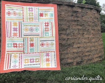 May include: A colorful patchwork quilt with a coral border is displayed outdoors. The quilt features various geometric patterns, including stars and rectangles, in pastel shades of pink, green, yellow, and blue. The quilt is hanging against a textured brick wall.