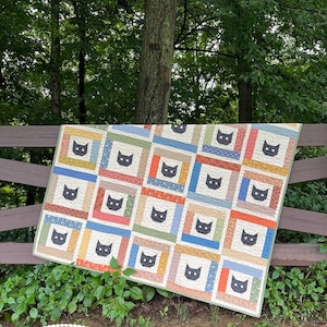 May include: A rectangular quilt featuring a repeating pattern of black cat faces centered within square frames. Each frame is constructed from various colorful fabric borders in shades of orange, blue, green, and tan, creating a grid layout.