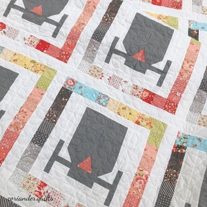 May include: Close-up of a handmade quilt with a repeating geometric design. The quilt features gray shapes with a red triangle accent, set against a white background. The design is framed by colorful patterned fabric strips.