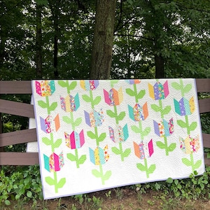 May include: A rectangular quilt featuring a repeating pattern of multi-coloured tulip flowers with bright green stems and leaves on a white background. The fabric is quilted with a curved, wave-like texture and finished with a light purple border.