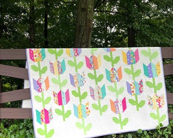 Op de afbeelding: Een rechthoekige quilt met een herhalend patroon van veelkleurige tulpenbloemen met felgroene stengels en bladeren op een witte achtergrond. De stof is doorgestikt met een golvende textuur en afgewerkt met een lichtpaarse rand.