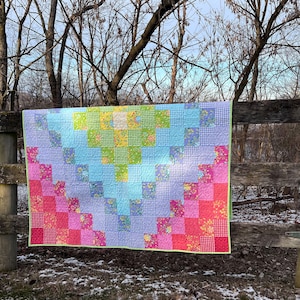 May include: A colorful patchwork quilt with a gradient design, transitioning from pink to blue and green. The quilt is made of various patterned squares and has a lime green border. It is draped over a wooden fence.