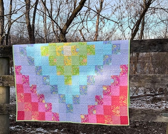 Op de afbeelding: Een kleurrijke patchworkquilt met een gradiëntontwerp, van roze naar blauw en groen. De quilt is gemaakt van verschillende patronen en heeft een limoengroene rand. Hij is over een houten hek gedrapeerd.