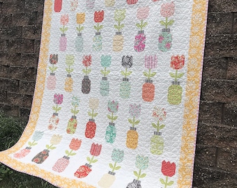 Op de afbeelding: Een handgemaakte quilt met een herhalend patroon van kleurrijke bloemvazen. De vazen zijn gevuld met diverse bloemmotieven en staan tegen een witte achtergrond. De quilt heeft een gele bloemenrand en de tekst "coriander.quilts" staat linksonder.
