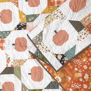 May include: A patchwork quilt featuring a repeating pumpkin design in shades of orange, white, and various fall-themed patterns. The quilt has a white background with orange pumpkin shapes and is bordered with a floral pattern. The quilt is made of fabric.