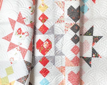 Op de afbeelding: Een close-up van een witte quilt met een verscheidenheid aan kleurrijke geometrische patronen. De quilt heeft rode, grijze, gele en blauwe ruit- en hartvormen, samen met stervormen. De quilt heeft een geschulpte rand en is gemaakt van zachte stof.