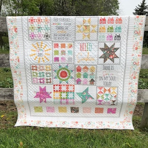 May include: A white patchwork quilt with various colorful squares, stars, and houses. The quilt features inspirational quotes like "KEEP YOUR FACE TOWARD THE" and "HELLO SUNSHINE." The edges are adorned with a floral pattern.