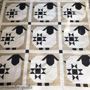 Puede incluir: Una colcha acolchada con un patr&oacute;n repetido de ovejas y estrellas. Las ovejas son blancas con caras y orejas negras, sobre una cuadr&iacute;cula beige y blanca. El texto "coriander quilts" es visible.