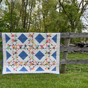 May include: A handmade quilt with a white background, featuring blue diamond shapes and colourful star patterns. The quilt is draped over a wooden fence in a grassy outdoor setting. The quilt's border has a floral pattern.