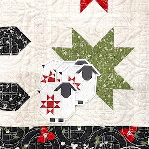 May include: A close-up of a quilt with a white background and a repeating pattern. The quilt features a green star, red bow, and black and white geometric shapes. Three sheep stickers are placed on the quilt. The quilt has the text "Calender Quilts by Corey Yoder".