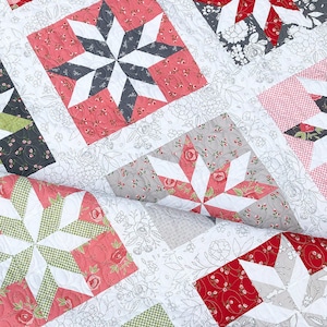 Peut inclure: Gros plan d'une courtepointe patchwork à motifs floraux. La courtepointe présente des motifs d'étoiles dans des tissus rouges, gris, blancs et verts. Le fond est blanc avec un motif floral subtil. Le texte "coriander quilts" est en bas à gauche.