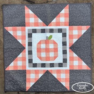 May include: A square quilt featuring a central pumpkin motif in orange and white gingham, surrounded by a black and white checkered border. A large eight-pointed star in orange gingham overlays a dark gray background with pumpkin quilting.