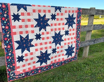 Könnte beinhalten: Eine handgefertigte Steppdecke mit patriotischem Thema, mit marineblauen Sternen auf weißem Hintergrund, durchsetzt mit roten Quadraten. Die Steppdecke hat einen floralen Rand in Marineblau, Rot und Rosa. Die Steppdecke hängt an einem Holzzaun.