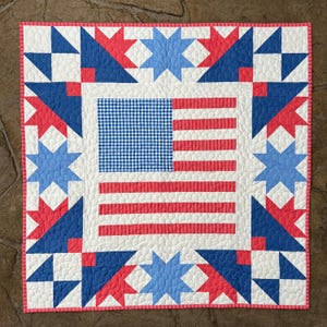 Könnte beinhalten: Ein quadratischer Quilt mit einem amerikanischen Flaggenmuster. Die Flagge hat ein blaues Vichy-Feld und rote und weiße Streifen. Der Rand hat blaue und rote Stern- und Dreieckmuster auf weißem Hintergrund, mit einem rosa Rand.