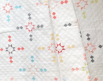 Op de afbeelding: Witte quilt met een herhalend patroon van kleine cirkels en kleurrijke ster- en ruitvormen. De sterren zijn rood, grijs, geel en roze. De ruiten zijn blauw, rood en grijs. De quilt is dubbelgevouwen.