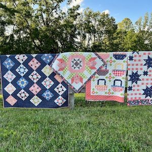 May include: Four handmade quilts displayed on a wooden fence. The quilts showcase diverse patterns, such as stars, diamonds, and floral motifs, using navy, pink, white, and red. The quilts are shown outdoors against a backdrop of green grass and trees.