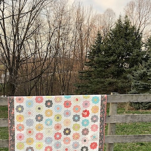 May include: A colourful floral quilt displayed on a wooden fence. The quilt has a white base with appliquéd flowers in pink, blue, yellow, and grey. The border is a dark floral pattern. The quilt is likely handmade.