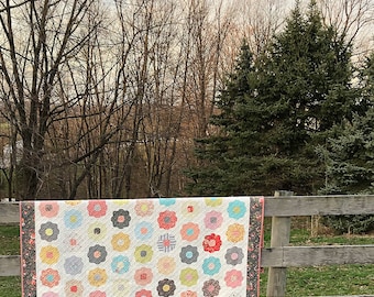Könnte beinhalten: Eine handgefertigte Steppdecke mit Blumenmuster, drapiert über einem Holzzaun. Die Steppdecke hat einen weißen Hintergrund mit bunten Blumenapplikationen in Rosa, Blau, Gelb und Grau. Der Rand ist ein dunkler Blumendruck.