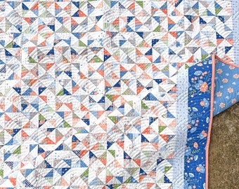 May include: A handmade quilt with a patchwork design. The quilt features a pattern of small triangles in shades of blue, coral, green, and gray on a white background. The border is blue with a floral pattern. The quilt is machine quilted.