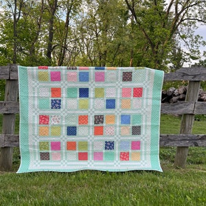 May include: A patchwork quilt with squares in shades of pink, blue, orange, and green, set against a light green border. The quilt is displayed on a wooden fence outdoors. The quilt is made by Coriander Quilts.