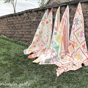 May include: Four colourful quilts leaning against a brick wall on green grass. The quilts feature various patterns, including stars and geometric shapes, in shades of red, yellow, green, and white. The text "coriander quilts" is visible at the bottom.