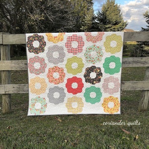 以下が含まれることがあります： 白い背景に、花柄やギンガムチェックなど、さまざまな模様と色の花型のブロックが特徴の手作りのキルト。キルトは木の柵に掛けられています。「coriander quilts」の文字が見えます。