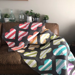 May include: A gray quilt with a pattern of colorful, striped hearts. The hearts are in shades of red, orange, yellow, green, blue, and purple. The quilt is draped over a brown leather couch. The text "coriander quilts" is visible.