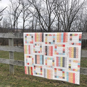 Puede incluir: Un edredón colorido con un diseño geométrico cuelga de una valla de madera. El edredón presenta un fondo blanco con bloques rectangulares en varios colores, incluyendo rosa, amarillo, gris y azul. El edredón se exhibe al aire libre con un telón de fondo de árboles.