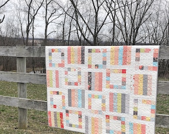 Può includere: Una colorata trapunta con un design geometrico è appesa a una staccionata di legno. La trapunta presenta uno sfondo bianco con blocchi rettangolari in vari colori, tra cui rosa, giallo, grigio e blu. La trapunta è esposta all'aperto con uno sfondo di alberi.