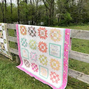 May include: A handmade quilt with a white background and colorful floral patterns, including pink, orange, and yellow. The quilt is hanging on a wooden fence outdoors, with a green border and a scalloped edge. The quilt has a message that says "KEEP YOUR FACE TOWARD THE SUNSHINE".