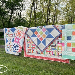 Könnte beinhalten: Mehrere bunte Quilts hängen an einem Holzzaun in einem grasbewachsenen Feld. Die Quilts weisen verschiedene geometrische Muster auf, darunter Sterne, Quadrate und Dreiecke, in Blau-, Orange-, Rosa- und Grüntönen. Die Quilts werden im Freien mit einem verschwommenen Hintergrund aus Bäumen ausgestellt.