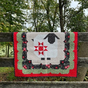 May include: A handmade quilt with a sheep motif, a red star, and floral details. The quilt showcases a white sheep with a black body on a beige backdrop, bordered by red, green, and black floral patterns. It's displayed on a wooden fence.