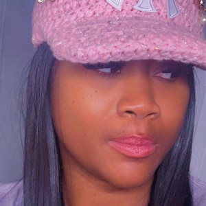 May include: Pink crocheted baseball cap with a silver and white logo. The cap features decorative embellishments, including a gold-colored pin with a flower design and small pearls. The cap has a curved brim and ear-like accents.