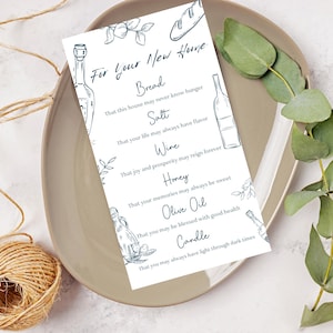 Puede incluir: Una tarjeta blanca con ilustraciones y texto en azul, presentada en un plato beige con eucalipto y cuerda. La tarjeta dice "Para tu nuevo hogar" y enumera pan, sal, vino, miel, aceite de oliva y vela.