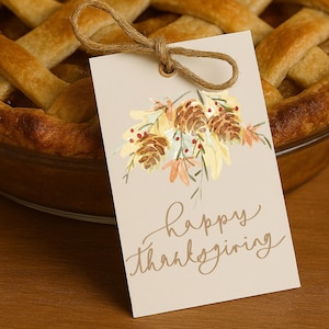 Può includere: Una torta al forno con una crosta dorata e un'etichetta decorativa. L'etichetta presenta un'illustrazione ad acquerello di pigne, foglie e bacche, con le parole "Happy Thanksgiving". È attaccato un fiocco di spago rustico.