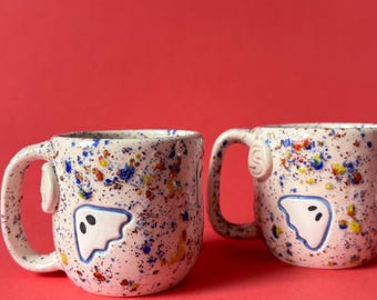 May include: Two ceramic mugs with a white base featuring a multi-colored paint splatter pattern in blue, yellow, and orange. Each mug displays a raised, white, ghost-shaped applique with two black eyes on the front and a small circular detail.