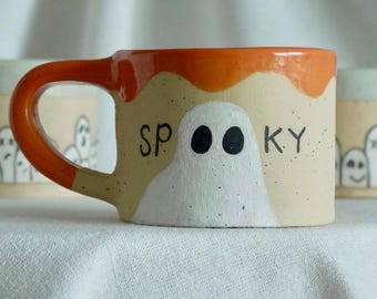 May include: A ceramic mug with a speckled beige finish, featuring a hand-painted white ghost and the black text "SPOOKY" on the front. The mug has a glossy orange rim and a matching orange handle, with a smooth, rounded cylindrical body shape.