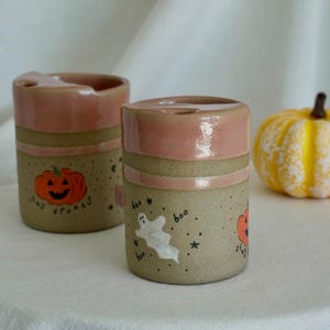 May include: Two cylindrical ceramic mugs with a smooth, dusty pink glazed rim and a speckled beige base. The mugs feature hand-painted Halloween motifs including an orange jack-o'-lantern with the text stay spooky and a white ghost with the text boo.