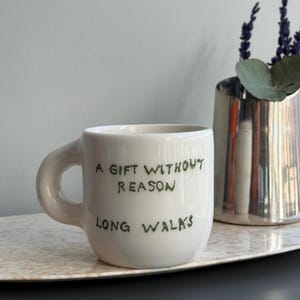 May include: Cream ceramic mug with a rounded handle, inscribed with the green text "A GIFT WITHOUT REASON LONG WALKS". A silver vase with lavender and greenery is in the background. The mug rests on a light-coloured tray.