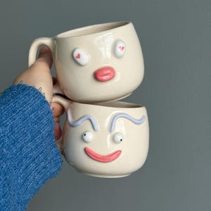 Könnte beinhalten: Zwei cremefarbene Keramikbecher, die übereinander gestapelt sind, jeder mit einem skurrilen Gesichtsdesign. Der obere Becher hat herzförmige Augen und einen rosa Mund. Der untere Becher zeigt ein lächelndes Gesicht mit blauen Augenbrauen und einem rosa Mund.